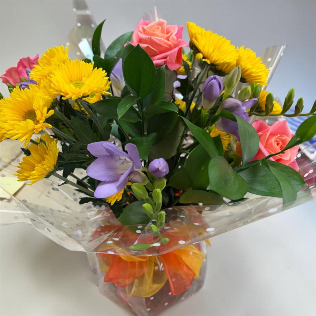 A bright hand-tied bouquet of yellow chrysanthemums, coral roses and lilac freesia in gift wrap, made by Bowbells Florist in Gloucester